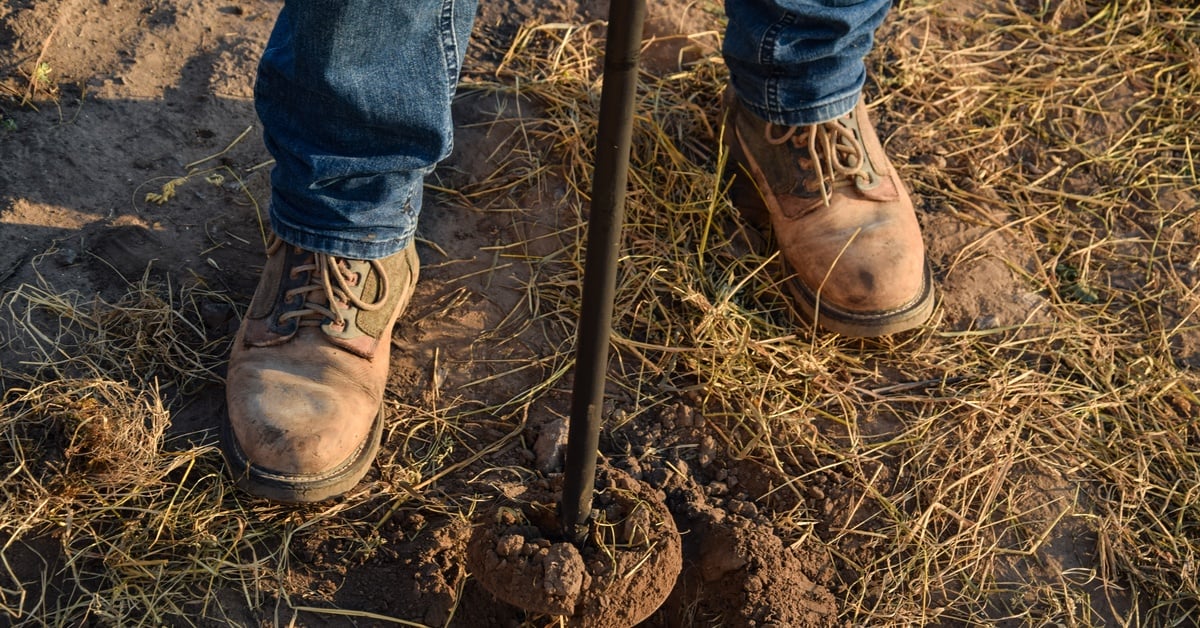 A close-up of a person wearing brown boots and jeans who is using a soil probe sampler on a plot of land.