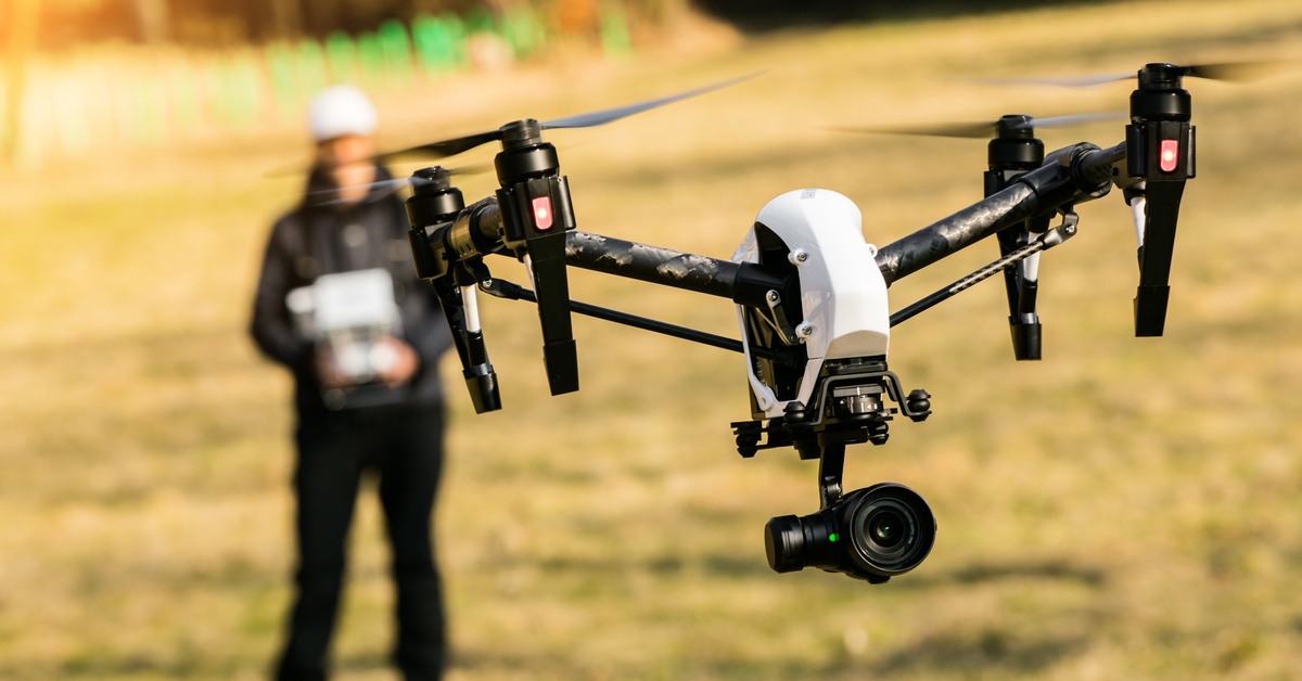 A black and white camera drone takes up the foreground. There is a drone operator with a controller in the background.