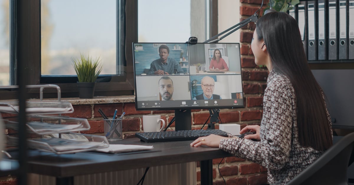 A remote worker looking at her desktop computer. The screen shows several other team members on a video call.