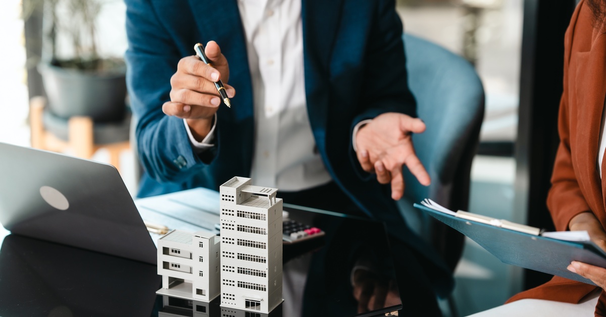 A man and woman talk over a laptop that is on the desk in front of them. The woman is holding a clipboard.