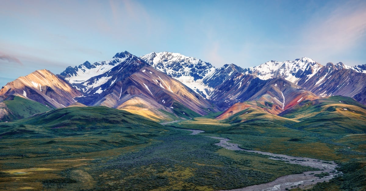 An aerial view of Denali National Park in Alaska. A wide river cuts through a forest leading to large mountains.
