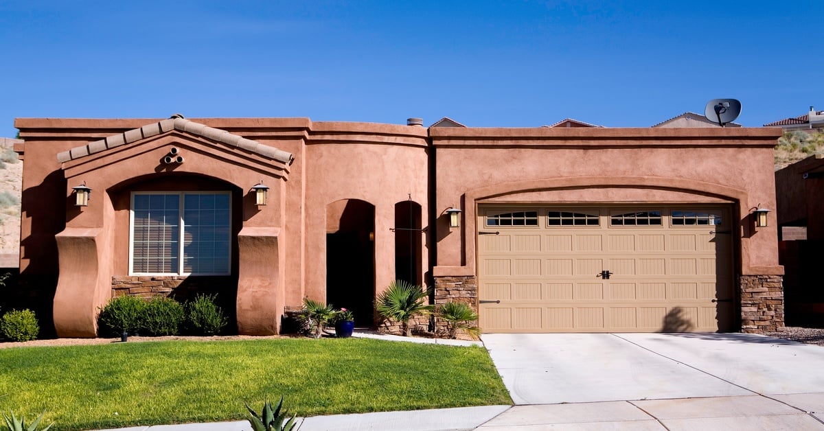A dark stone, Adobe-style house with an ornate entrance, a green grass lawn, and a white stone driveway.
