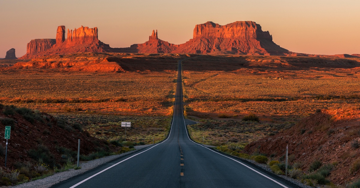 A long, straight road cutting through a desert landscape with large plateaus in the distance during a sunrise.