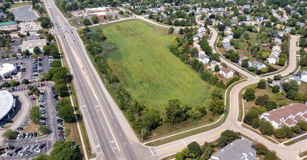 An aerial shot of a grassy area in the middle of a neighborhood. Houses and streets surround the empty lot.