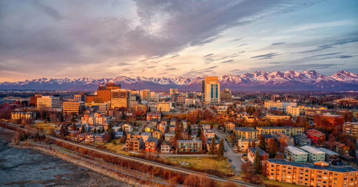 A distant aerial view of the skyline of Anchorage, Alaska. The sun is setting behind the various buildings.
