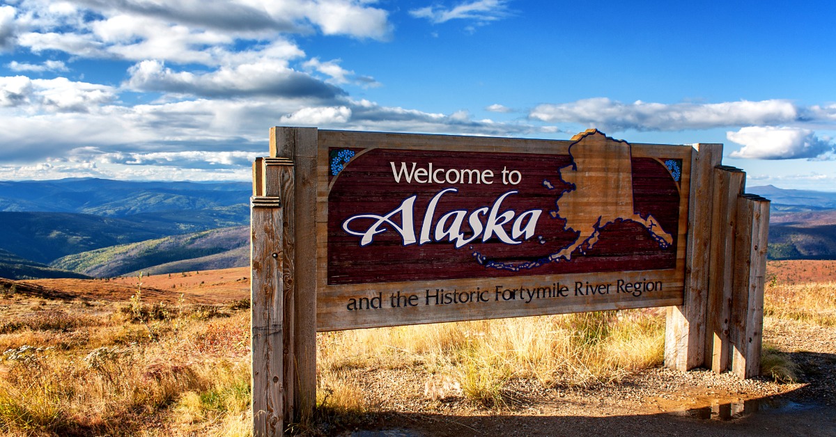 A wide wooden sign that says "Welcome to Alaska" across it. There are many grassy hills stretching behind the sign.