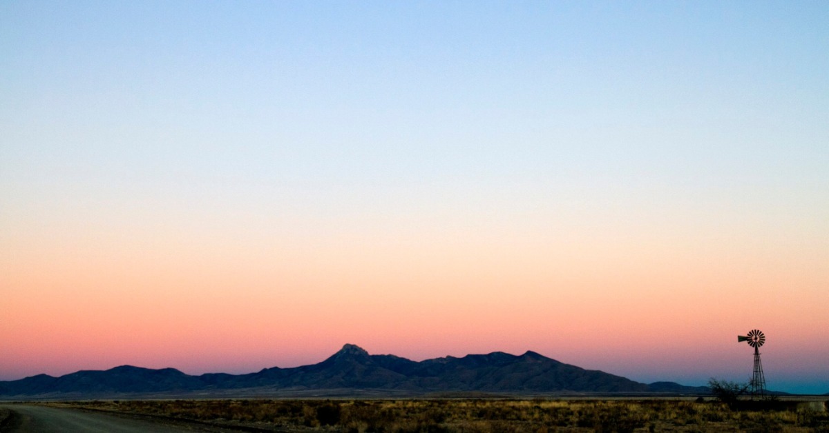 A wide shot of a small mountain range against the sky at sunset. There is a small windmill on the plains near the mountains.