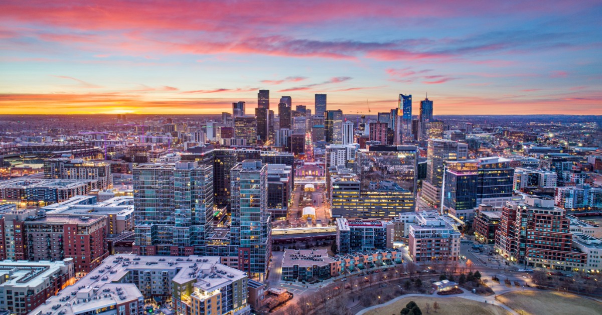 An aerial drone shot of the skyline of Denver, Colorado. The buildings are set against a sky at sunset.
