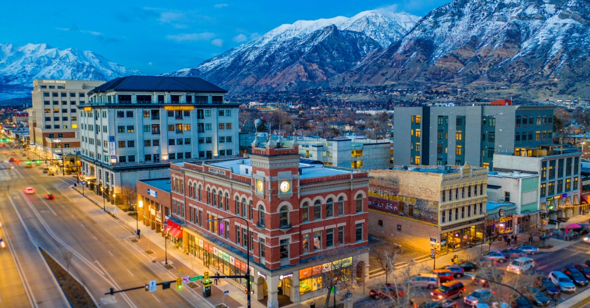 The buildings that make up the downtown section of Provo, Utah. Tall mountains stretch upwards from behind the buildings.