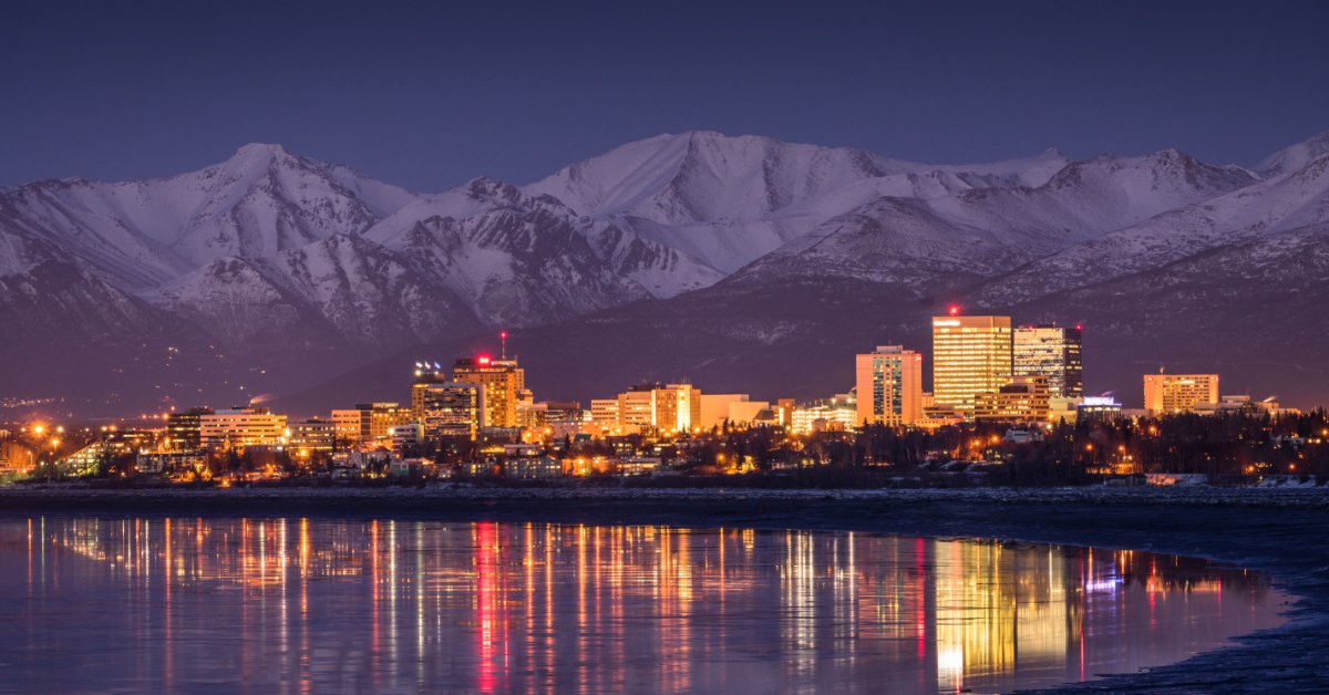 A far view of the skyline of Anchorage, Alaska at night. A large mountain range is just behind the skyline.