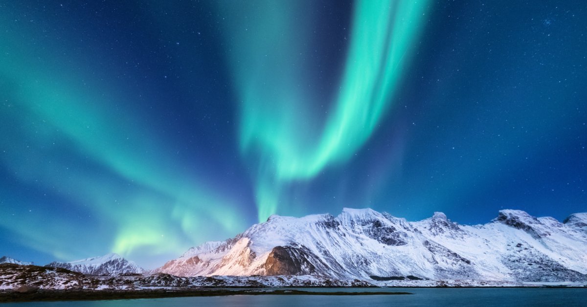 The Aurora Borealis lights shining green and yellow above a large, snow-capped mountain range and a lake.