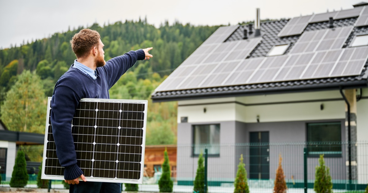 A man holding a solar panel pointing up at a house's roof that also is covered in several solar panels.