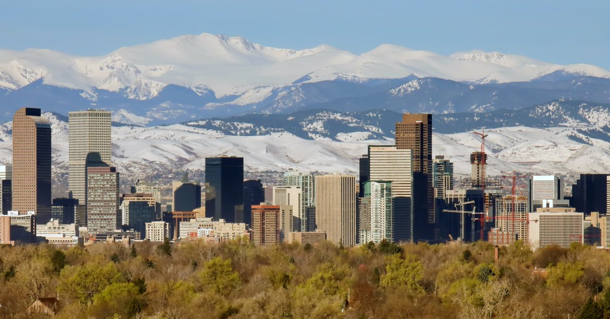 A distant view of the skyline of Denver, Colorado. There are large mountains in the distance behind the buildings.