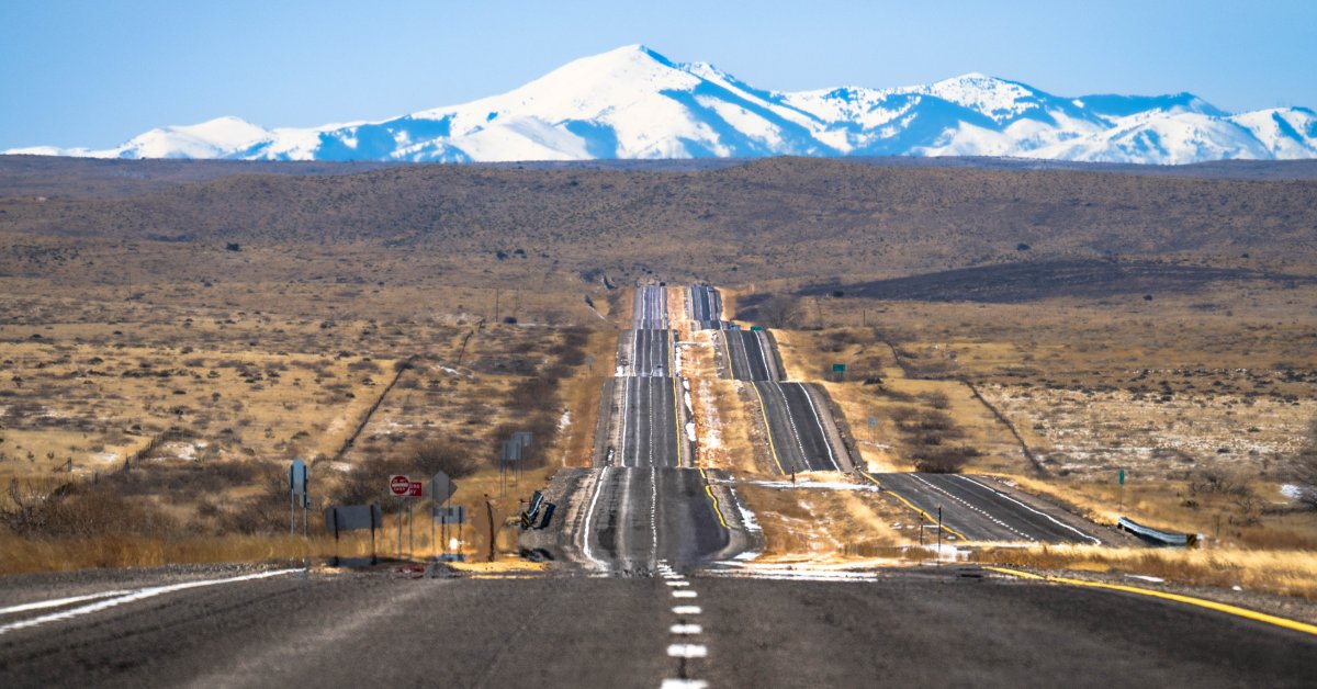 A view down a long road that cuts through some plains. There are large, snowcapped mountains in the background.