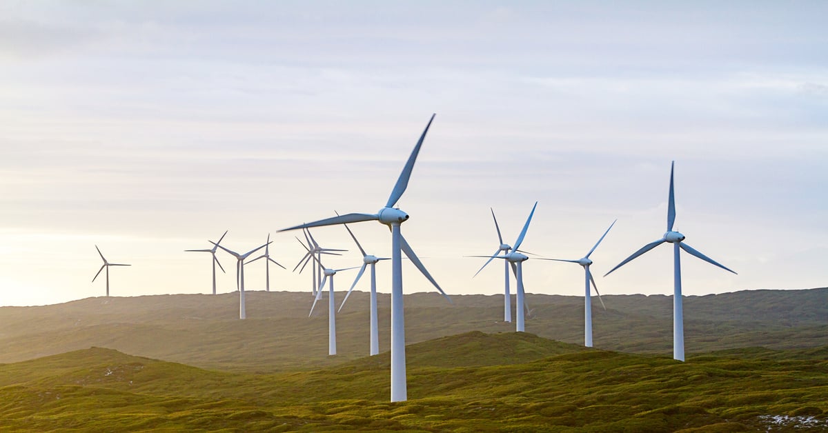 Large plot of land covered in green grass and small hills with multiple white wind towers scattered around the area.