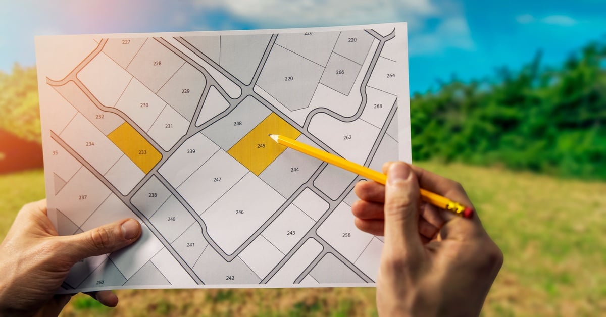 A hand holding up a map with land divisions. The person's other hand uses a pencil to point at a plot colored in yellow.