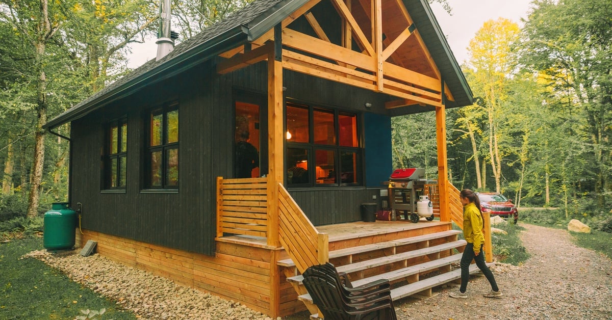 A one-story wooden cabin with a pointed roof and a small patio in a forest. A girl wearing a yellow coat is walking toward it.