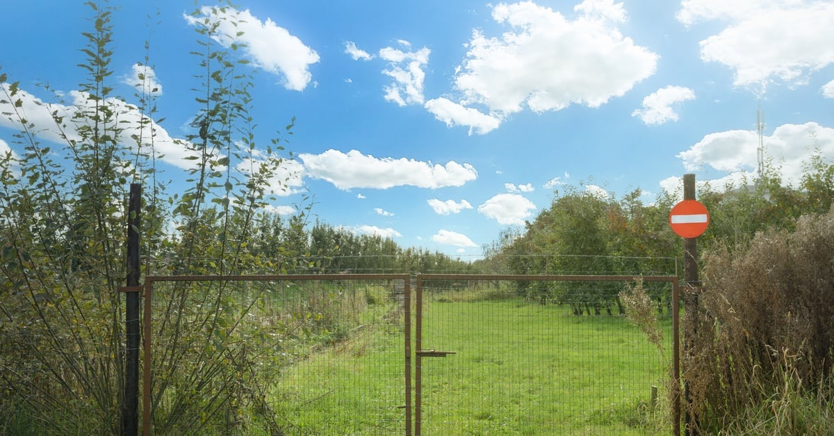 A closed gate in front of a grassy lot with a prohibited entrance sign. The land is overgrown, and the sky is bright blue.