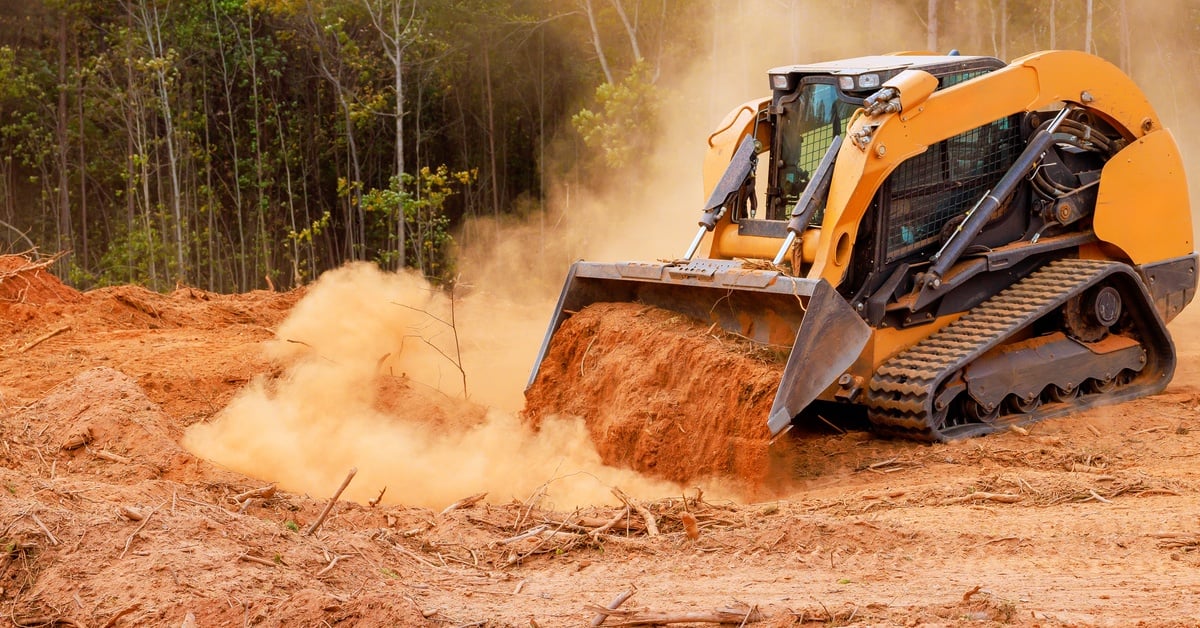 A small bulldozer is moving soil inside a large plot of land with multiple thin trees in the background on a sunny day.