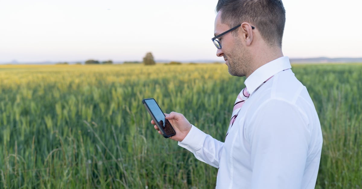 A man wearing a white shirt holds a smartphone in his right hand while standing on a large plot of land.