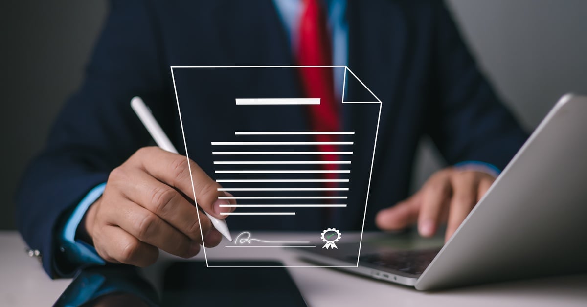 A person in a navy suit sits at a desk with a laptop and writes on a tablet with a document graphic superimposed over it.