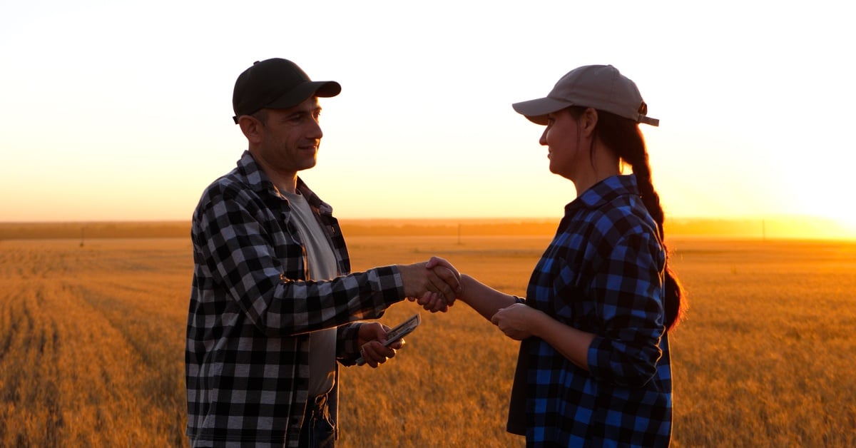 A man and woman wearing hats and plaid shirts shake hands while standing on a large plot of land at sunset.