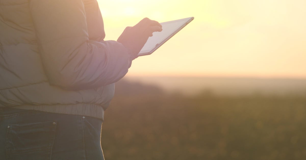 A close-up of a person's holding a tablet while standing on a plot of land during a sunset. The person wears a jacket.