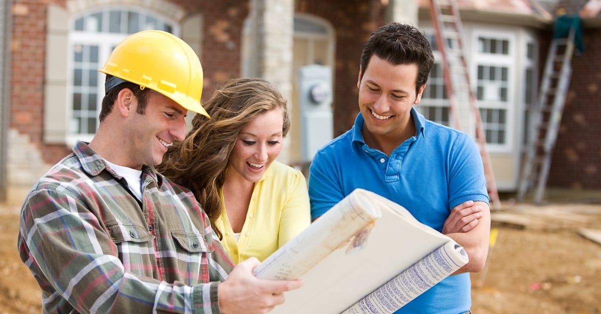 A contractor wearing a yellow helmet, showing a man and a woman a large paper plan in front of a house under construction.
