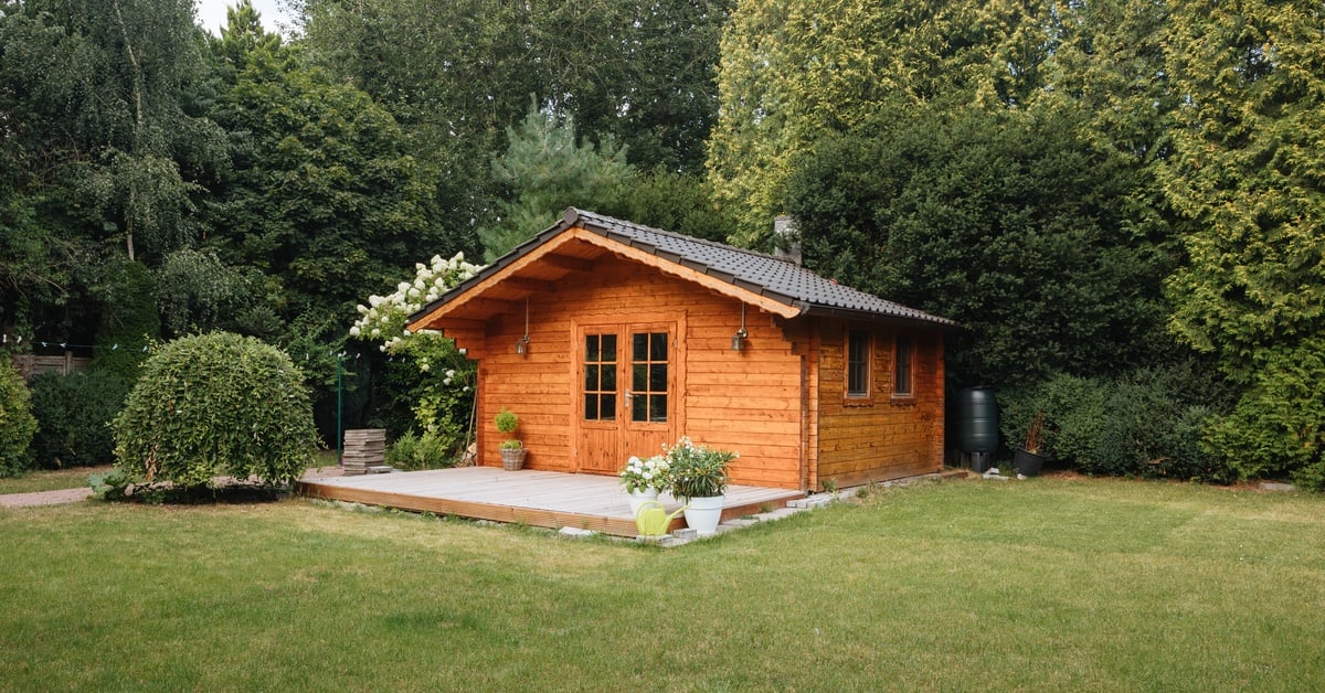 Small wooden cabin with a roof covered in brown tiles, placed over lush green grass, surrounded by trees and bushes.