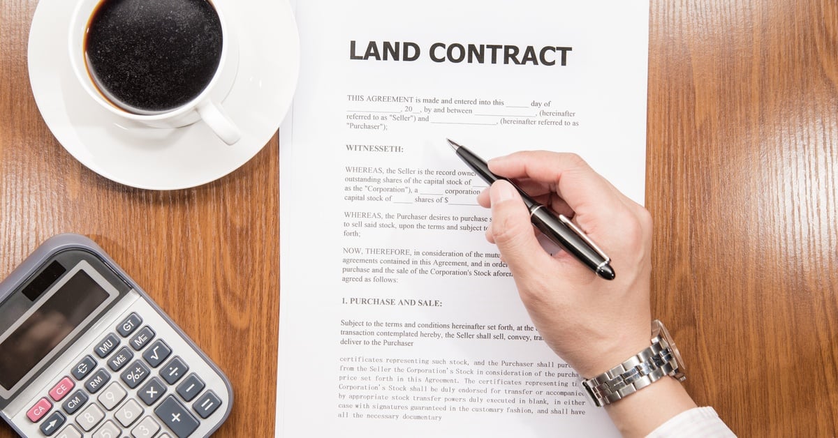 Aerial view of a wooden desk with a coffee mug, a calculator, and a hand signing a paper displaying “land contract”.
