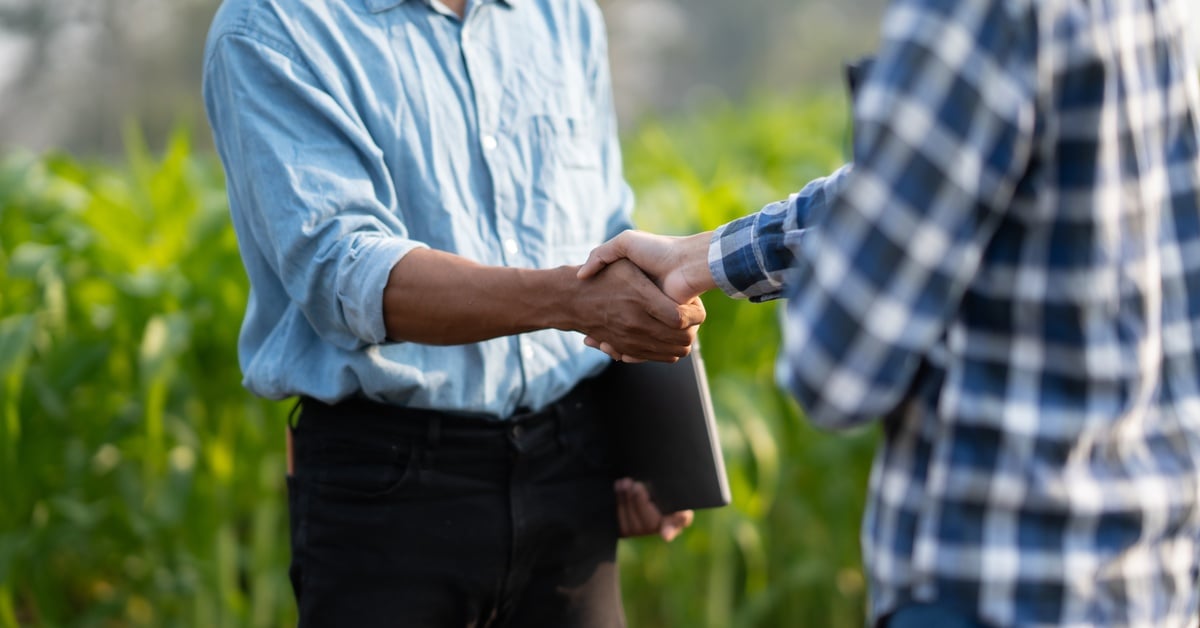 Man wearing a plaid shirt shaking the hand of a man holding a table while standing in front of green crops.