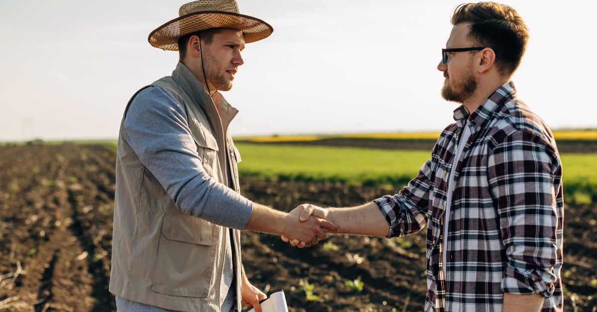 A man wearing a sunhat and a brown vest shaking the hand of a man wearing a plaid shirt while standing on farmland.