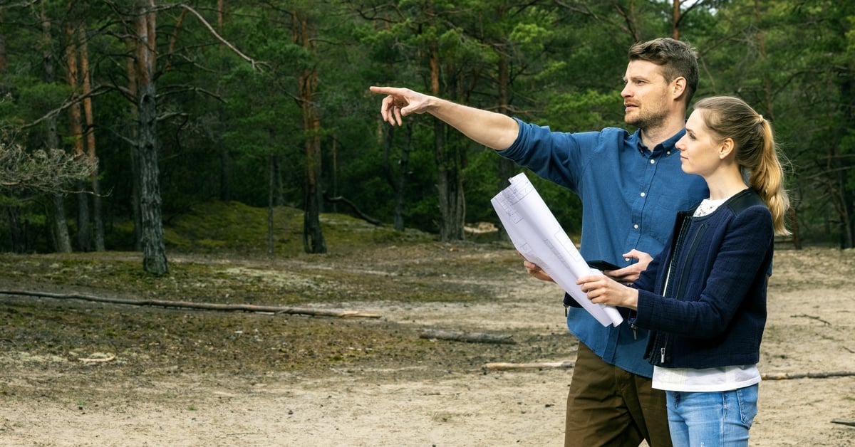 A man standing with a woman in a wooded area. The man raises an arm and points while a woman holds a large paper.